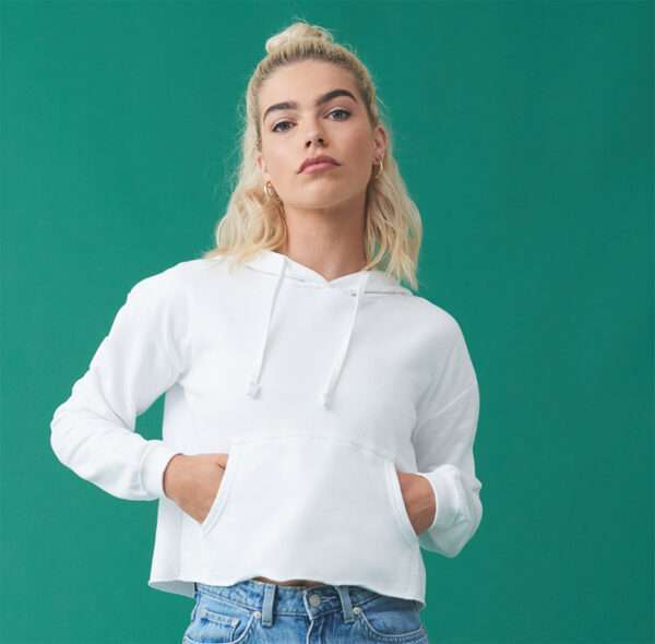 Junge Frau mit blondem Dutt in weißem Hoodie, Hände in Kängurutasche, grüner Hintergrund